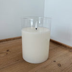 White candle in a clear glass container on a wooden surface with a white wall background