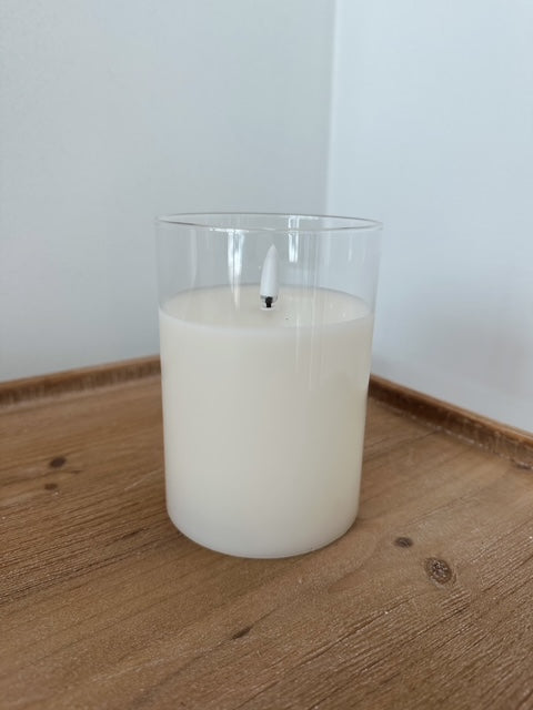 White candle in a clear glass container on a wooden surface with a white wall background
