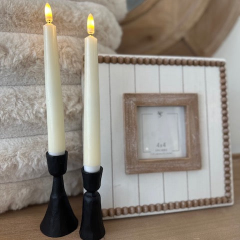 Two lit candles in black holders on a wooden surface with a framed picture in the background.