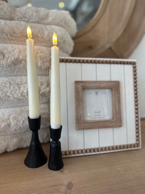 Two lit candles in black holders on a wooden surface with a framed picture in the background.