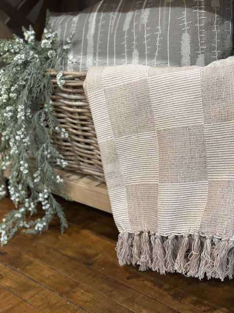 Textured blanket with fringe draped over a wicker chair on a wooden floor.