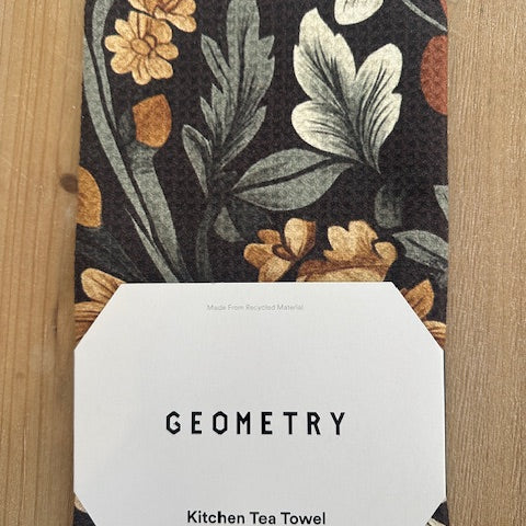 Floral patterned kitchen tea towel with a white tag displaying 'GEOMETRY' on a wooden surface.