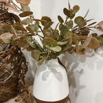 White vase with brown base holding green leaves on a wooden surface