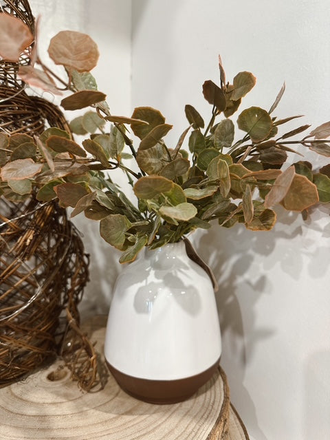 White vase with brown base holding green leaves on a wooden surface