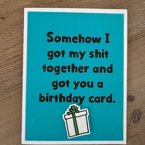 Blue birthday card with humorous text and a gift illustration on a wooden surface