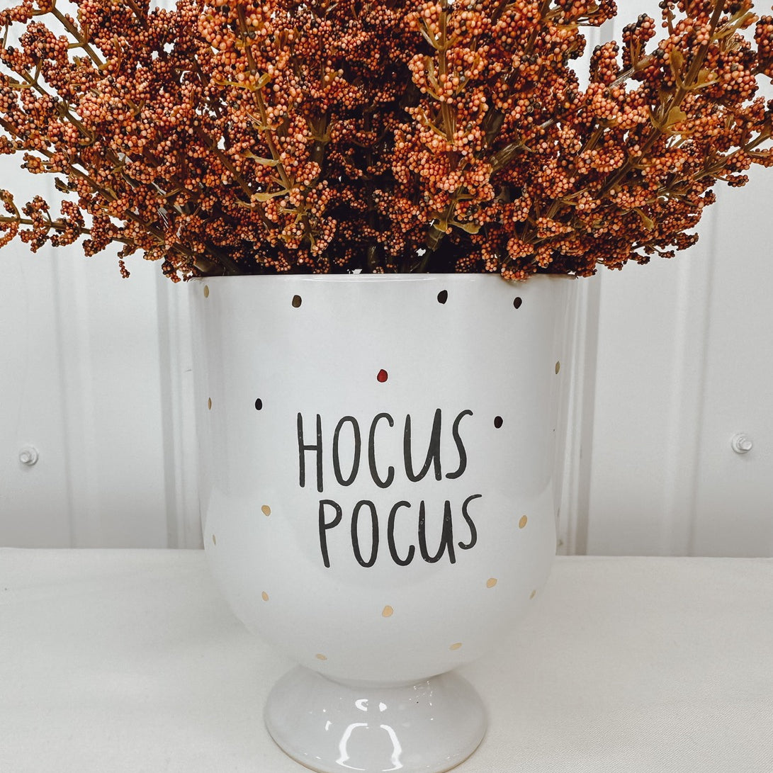 A white ceramic planter with the phrase 'Hocus Pocus' in black text, surrounded by brown artificial flowers.