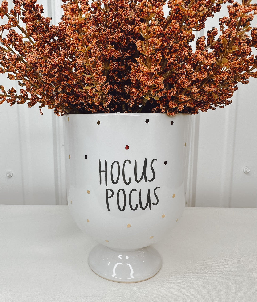 A white ceramic planter with the phrase 'Hocus Pocus' in black text, surrounded by brown artificial flowers.