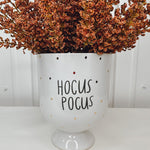A white ceramic planter with the phrase 'Hocus Pocus' in black text, surrounded by brown artificial flowers.